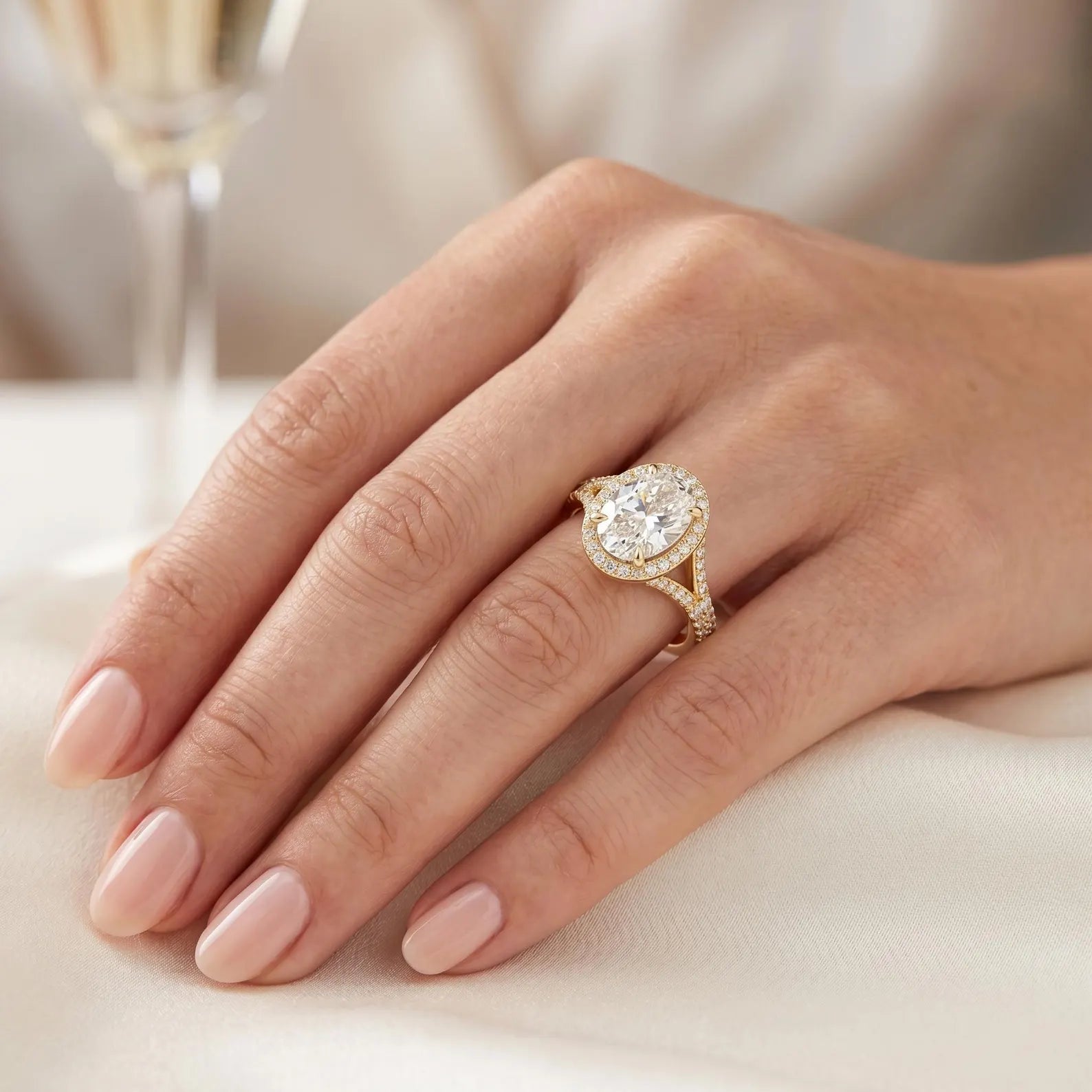 Close-up perspective on hand showing a 3ct oval lab diamond split-shank halo engagement ring in yellow gold.