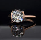 Front view of a 3 carat cushion cut lab grown diamond set in a full gold bezel ring on a black reflective background.