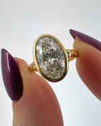 Close-up shot of a 4 carat oval lab grown diamond milgrain bezel ring held between fingers showing brilliant sparkle
