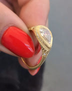 Hand holding a 2 carat pear lab grown diamond ring with milgrain bezel and flush-set side diamond accent in yellow gold