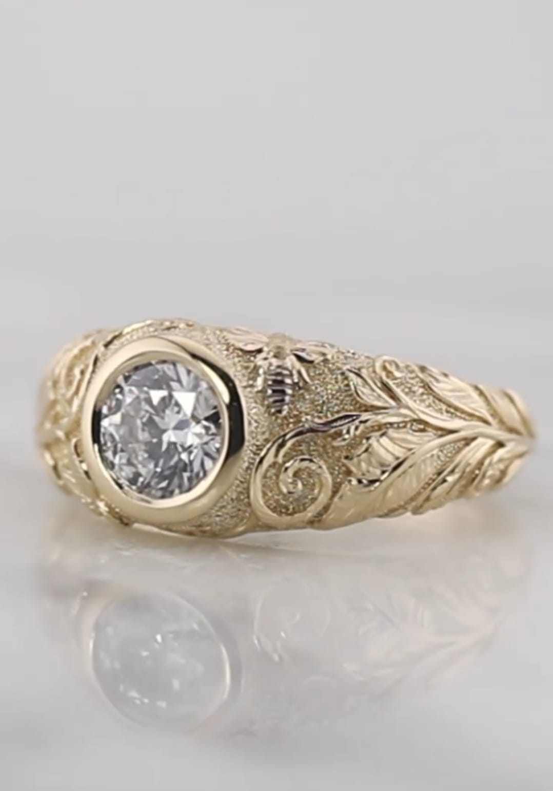 Angled studio shot of a 2 carat round lab grown diamond bezel ring featuring hand-engraved leaf patterns and a bee motif on yellow gold
