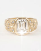 Front-facing shot of a vintage-inspired yellow gold ring showcasing a clear emerald-cut diamond center stone in a bezel setting, flanked by an intricately detailed, textured wide band with inset round diamonds.