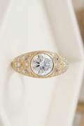 Close-up profile view of a vintage-style gold engagement ring featuring an intricate filigree band, round-cut central diamond moissanite, and detailed baguette accents.