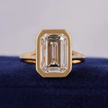 Front view of a 3 carat emerald cut lab grown diamond engagement ring in a yellow gold bezel setting resting on blue velvet.
