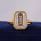 Front view of a 3 carat emerald cut lab grown diamond engagement ring in a yellow gold bezel setting resting on blue velvet.