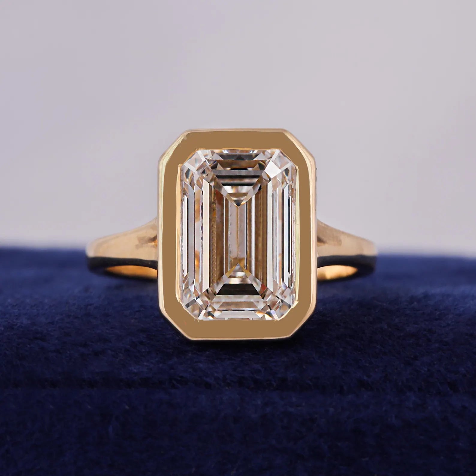 Front view of a 3 carat emerald cut lab grown diamond engagement ring in a yellow gold bezel setting resting on blue velvet.