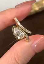 Close-up held view of a 1.5 CT pear cut lab grown diamond bezel ring with a pavé diamond bypass band in yellow gold.