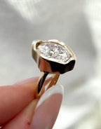 A close-up of a gold ring with a large, irregularly shaped clear gemstone held between fingers against a soft