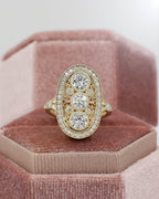 Vintage-style elongated oval three-stone diamond ring in yellow gold with filigree and milgrain detailing, resting inside a pink velvet hexagon ring box.