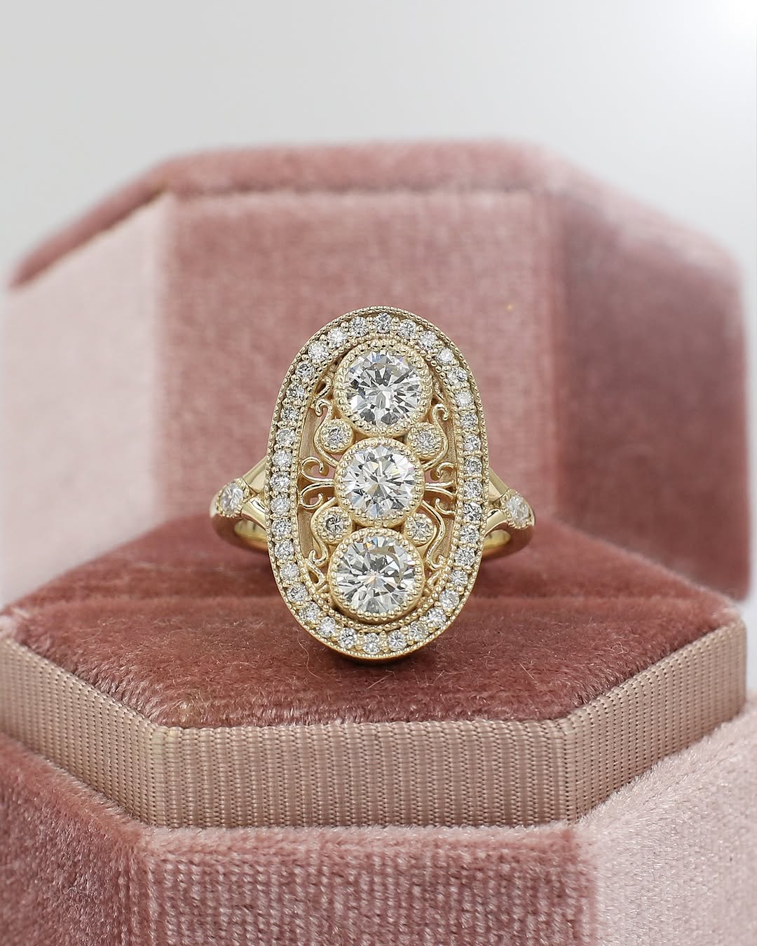 Vintage-style elongated oval three-stone diamond ring in yellow gold with filigree and milgrain detailing, resting inside a pink velvet hexagon ring box.