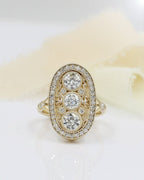 Top-down view of an Art Deco style diamond ring on a white surface with cream ribbon, featuring three round center stones, filigree swirls, and a diamond halo in yellow gold.