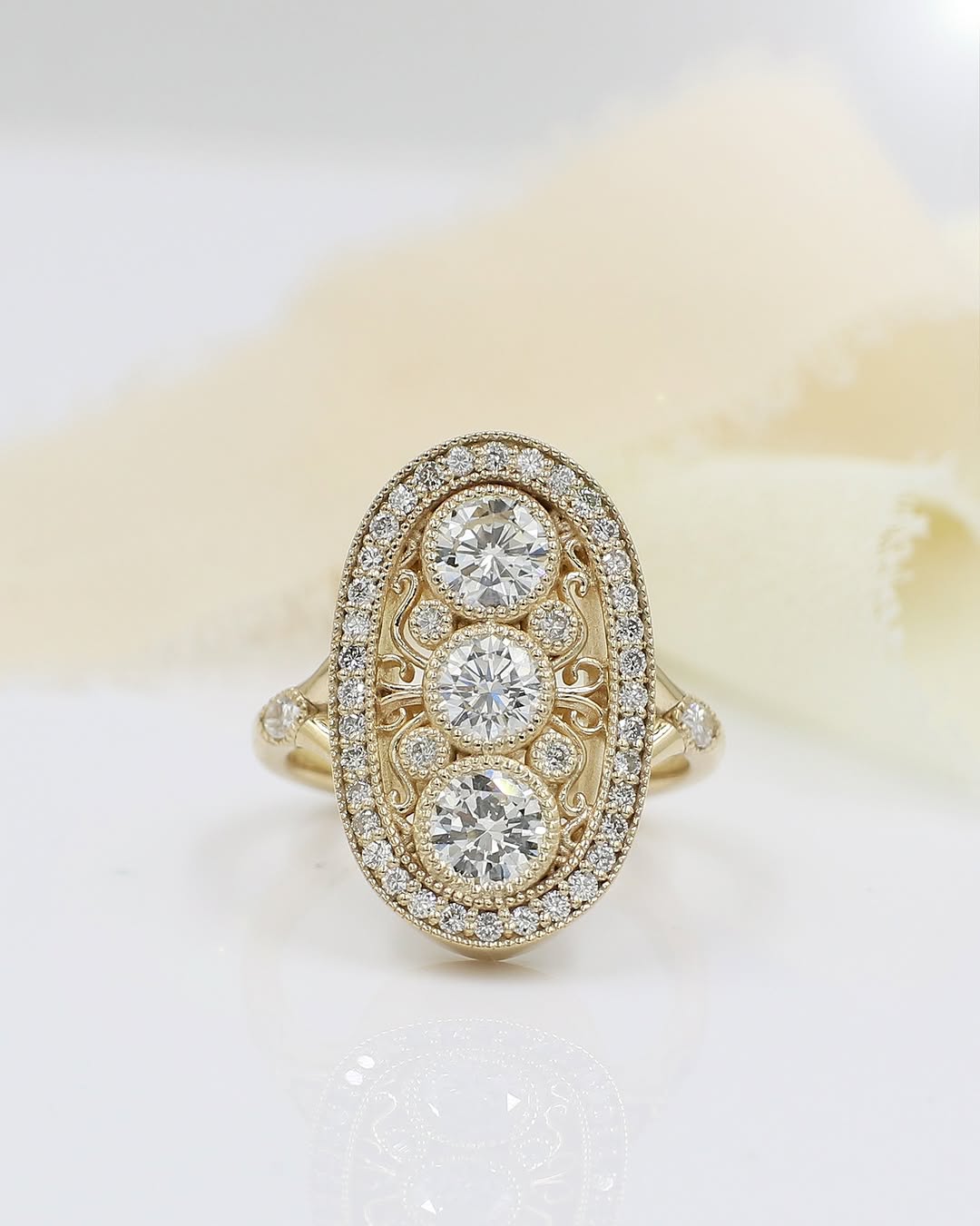 Top-down view of an Art Deco style diamond ring on a white surface with cream ribbon, featuring three round center stones, filigree swirls, and a diamond halo in yellow gold.