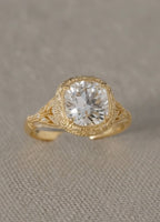 Top-down view of a vintage-inspired cushion-shaped ring in yellow gold resting on a textured fabric surface, highlighting the elaborate metalwork and center stone.