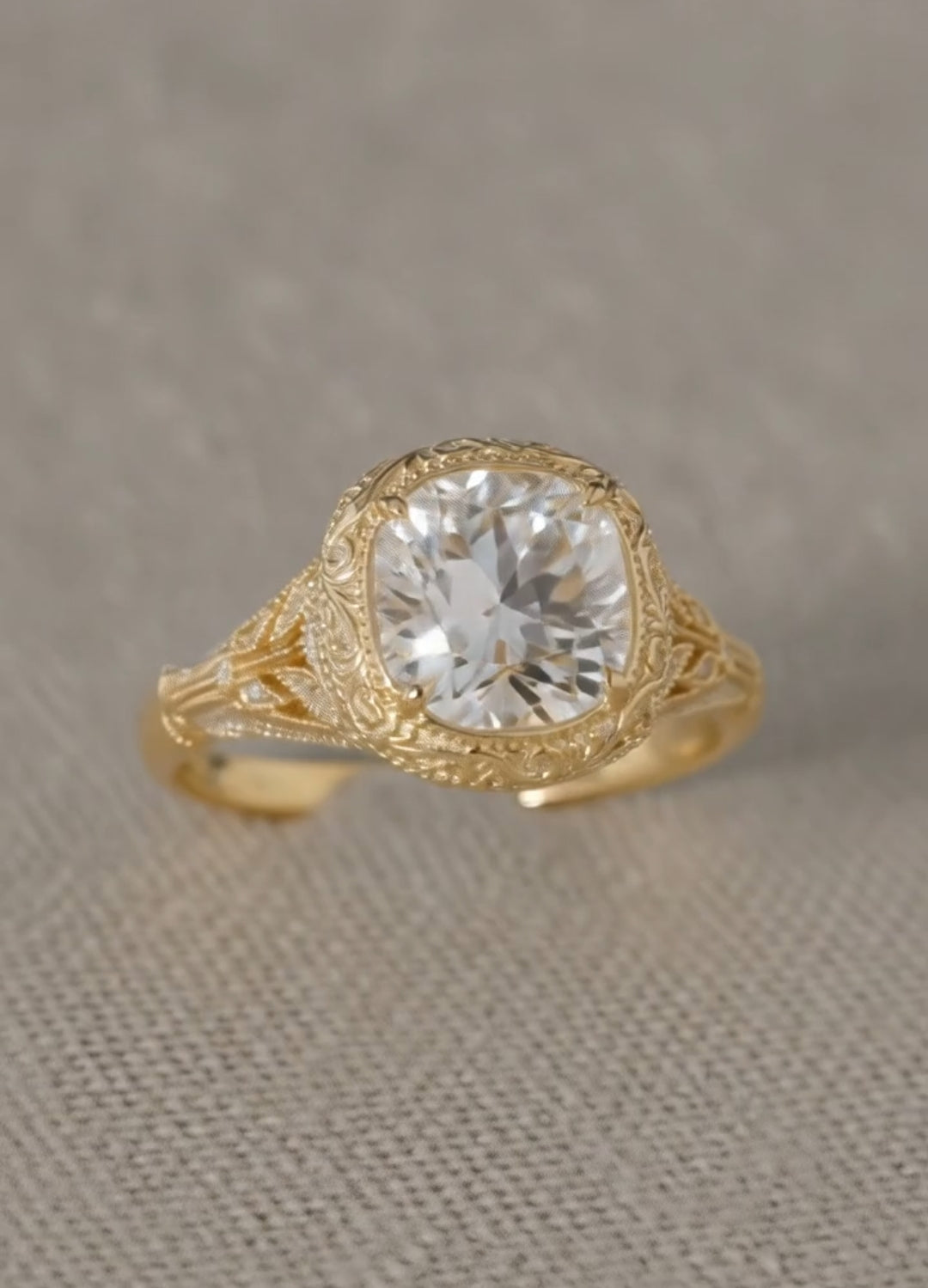 Top-down view of a vintage-inspired cushion-shaped ring in yellow gold resting on a textured fabric surface, highlighting the elaborate metalwork and center stone.