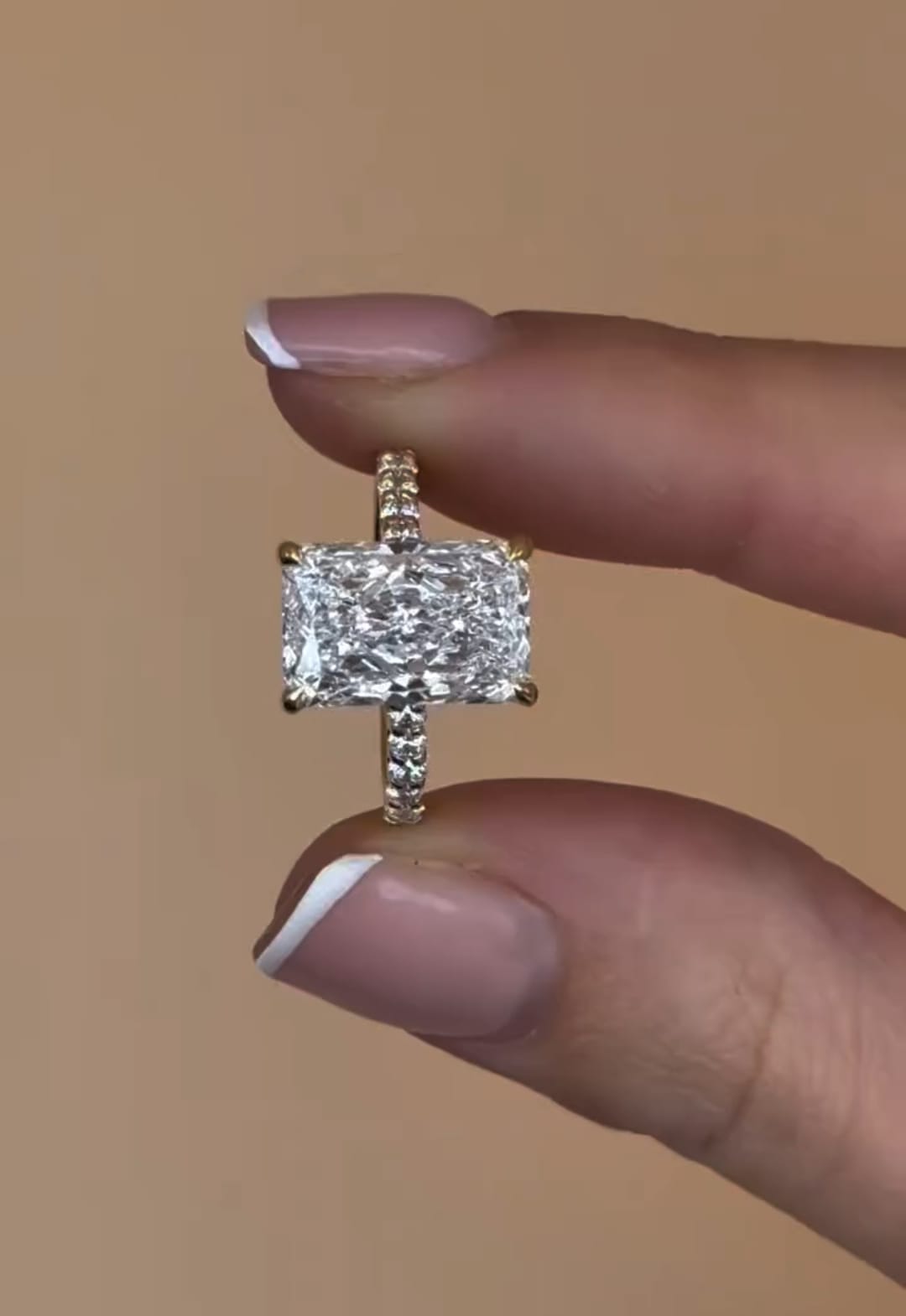 Close up front view of a 3 carat radiant cut lab diamond engagement ring held between two fingers, highlighting its brilliant facets.