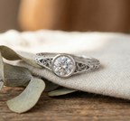 Close-up top view of a vintage-style white metal engagement ring featuring a round center stone in a milgrain bezel setting with filigree shoulders.