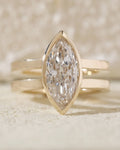 Close-up front view of a brilliant elongated lab grown diamond in a gold bezel setting resting on a textured stone surface.