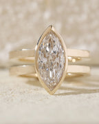 Close-up front view of a brilliant elongated lab grown diamond in a gold bezel setting resting on a textured stone surface.