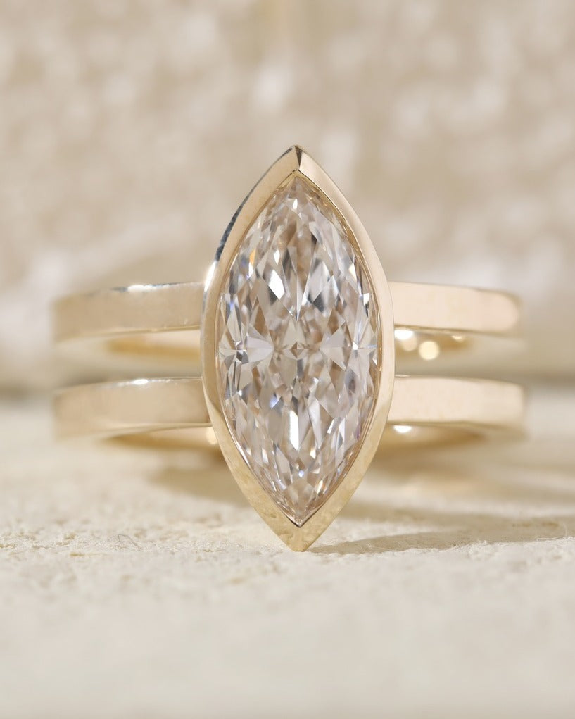 Close-up front view of a brilliant elongated lab grown diamond in a gold bezel setting resting on a textured stone surface.