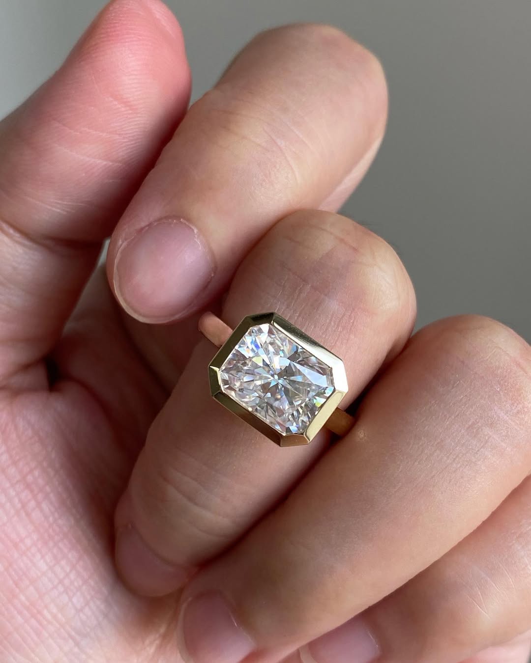 On-finger view of a sleek east-west bezel set 3 CT radiant cut lab diamond engagement ring in yellow gold.