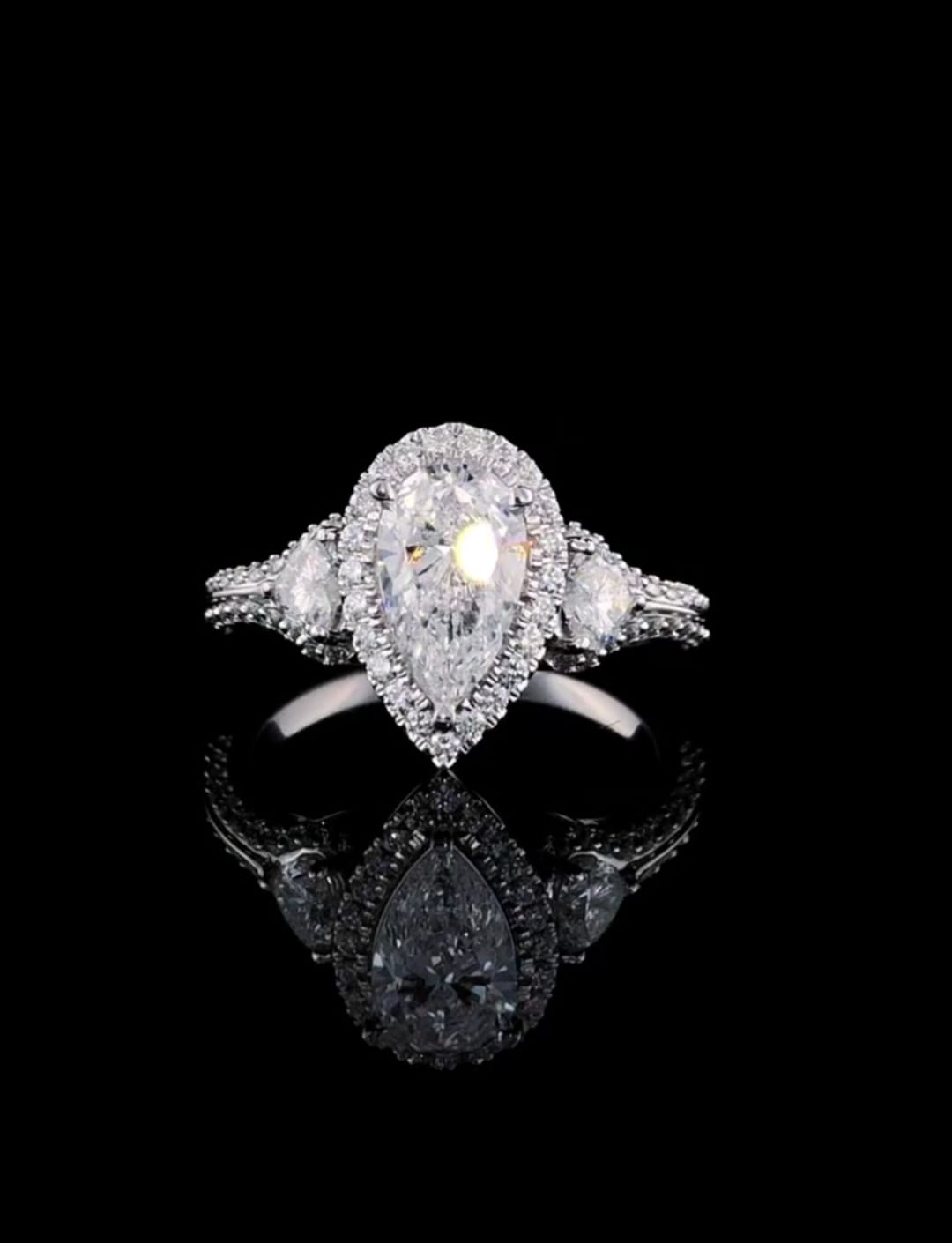 Front view of a 3 CT pear cut lab grown diamond engagement ring with a halo and trillion side stones on a black background
