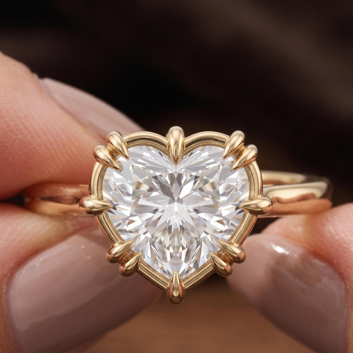 A heart-shaped diamond ring in a gold prong setting, held between two fingers against a dark, blurred background.
