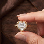 A close-up of a hand holding a gold ring with a heart-shaped clear gemstone, set against a dark brown textured background.