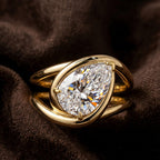 A gold ring featuring a large pear-cut clear gemstone in a prong setting, resting on a dark brown textured fabric background.