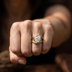 A close-up of a hand wearing a gold ring with a pear-cut clear gemstone. The ring is shown on a finger against a dark, softly blurred background.