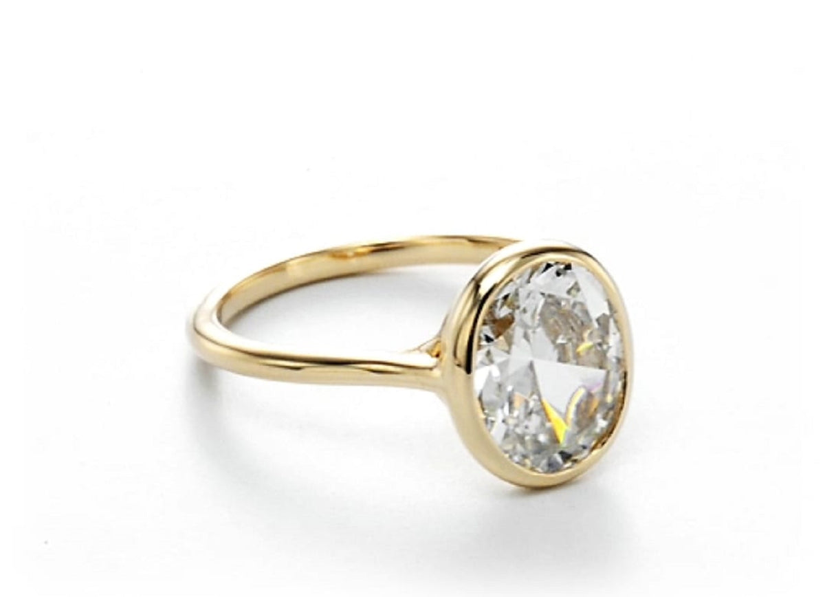 Side angle view showcasing the slim polished gold band and oval lab diamond bezel ring on white background.