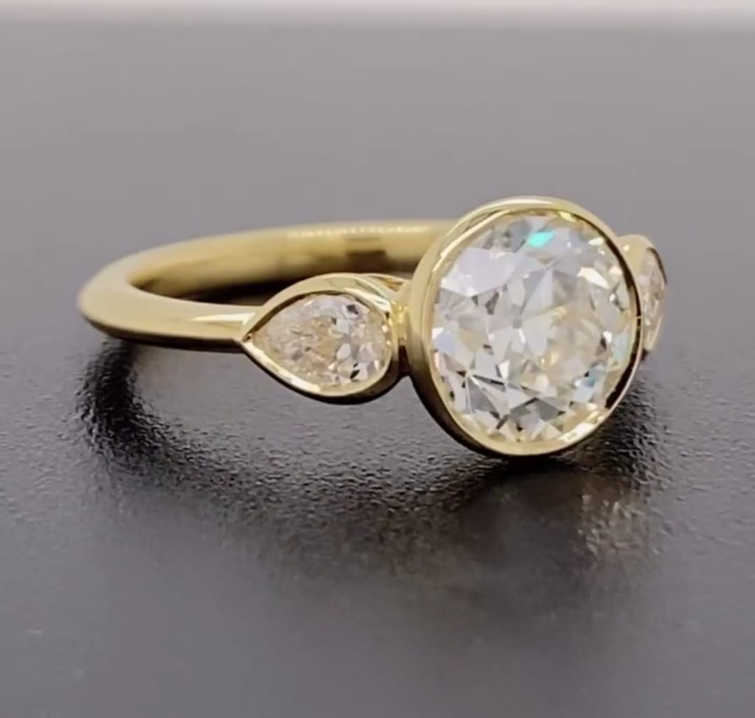 Side profile of a 3 carat round lab grown diamond bezel engagement ring with pear side stones in yellow gold.