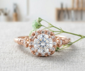 Close-up of a vintage-style rose gold engagement ring featuring a round center stone and detailed halo, resting on a textured fabric background.