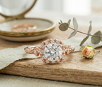 Rose gold engagement ring with a detailed halo setting and milgrain band, resting elegantly on a wooden surface next to a small dried rosebud