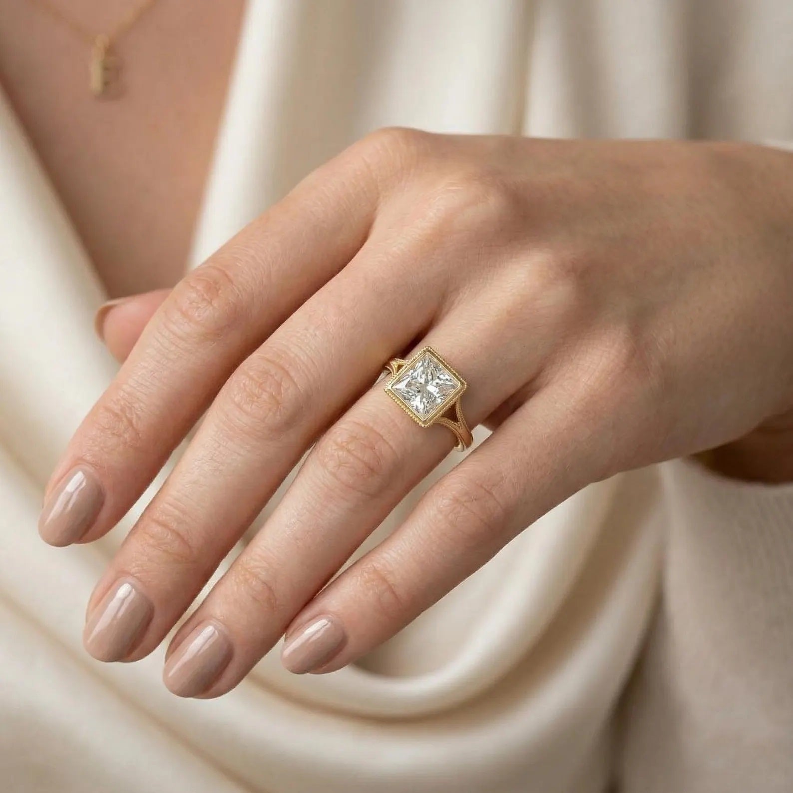 A woman wearing a vintage-style yellow gold engagement ring with a bezel-set square center stone and split shank, resting her hand elegantly against a cream-colored silk garment.