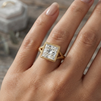 Close-up of a hand wearing a yellow gold Art Deco-style engagement ring featuring a princess-cut center stone, milgrain bezel setting, and a detailed split shank.