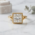 Front macro view of a dazzling princess-cut bezel set yellow gold engagement ring with intricate milgrain detailing, resting on white marble with a blurred velvet ring box in the background