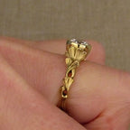 A gold ring with a decorative leaf-like design and a clear gemstone, shown worn on a person's finger against a neutral beige background.