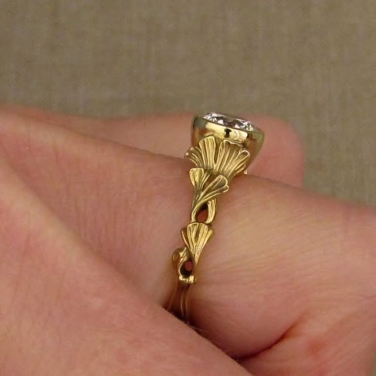 A gold ring with a decorative leaf-like design and a clear gemstone, shown worn on a person's finger against a neutral beige background.