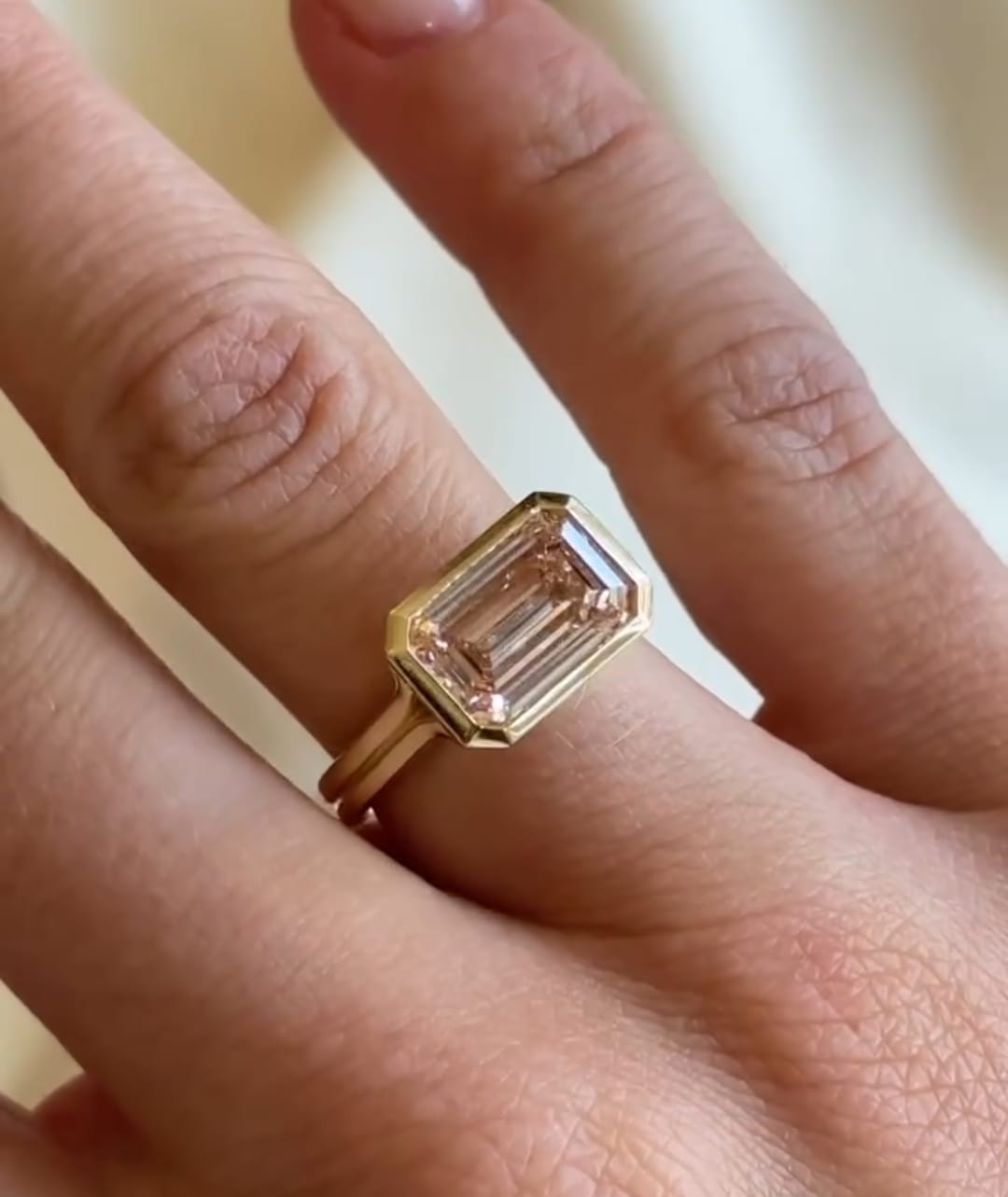 3 carat blush pink emerald cut lab created diamond engagement ring worn on finger in yellow gold bezel setting.