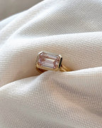 3 CT pink emerald cut lab grown diamond ring with yellow gold bezel setting displayed on white fabric.