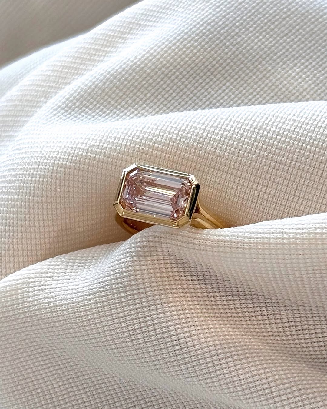 3 CT pink emerald cut lab grown diamond ring with yellow gold bezel setting displayed on white fabric.