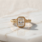 Front view of a 3ct elongated radiant cut lab grown diamond engagement ring with a milgrain bezel and engraved band resting on white marble.