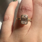 Close-up on hand showing a 3 CT emerald cut lab grown diamond bezel engagement ring with engraved yellow gold band.