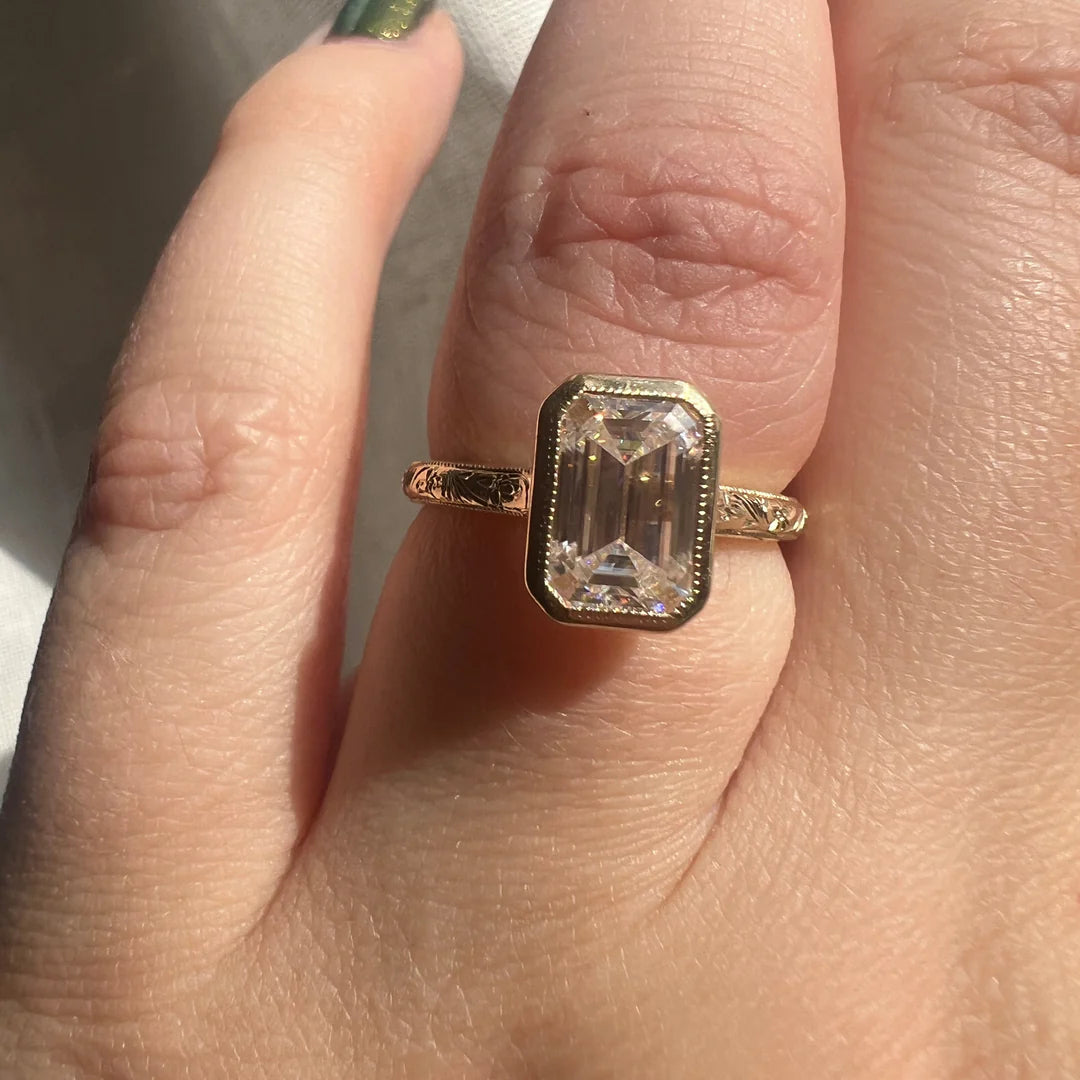 Close-up on hand showing a 3 CT emerald cut lab grown diamond bezel engagement ring with engraved yellow gold band.