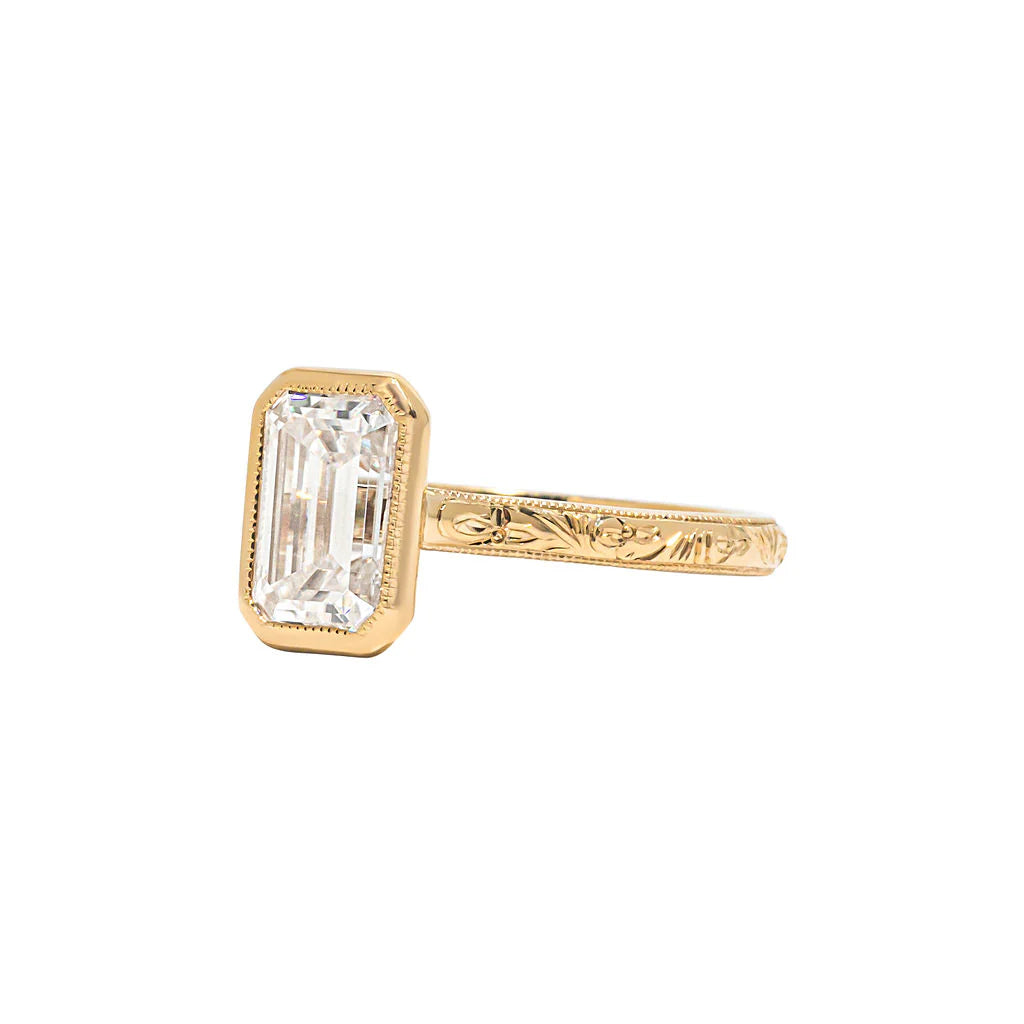 Angled studio view of 3 CT emerald cut lab diamond bezel ring highlighting intricate floral scroll engraved band.