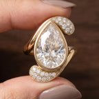 Close up of yellow gold 3 CT pear cut lab diamond bezel ring held in fingers.