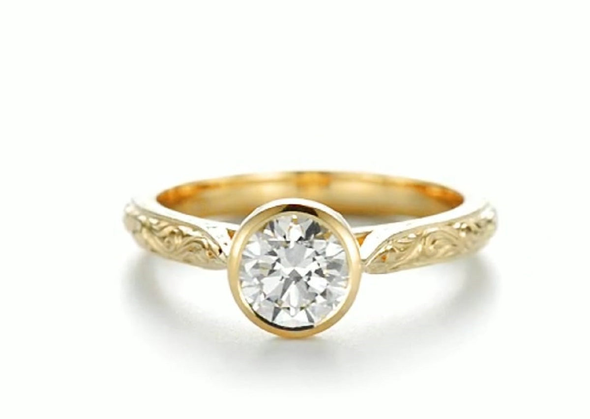 Front view of a 2 CT round lab grown diamond bezel set engagement ring in hand-engraved 18K yellow gold.
