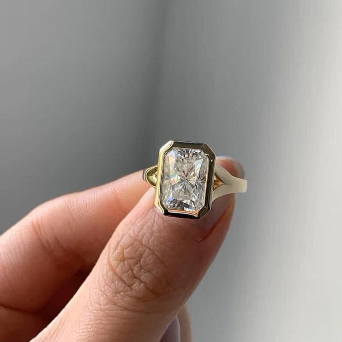 Hand holding a modern 4 CT radiant cut lab diamond solitaire ring with smooth yellow gold bezel setting.