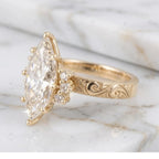 Close-up profile of a yellow gold engraved marquise engagement ring showing side diamond cluster accents on marble.