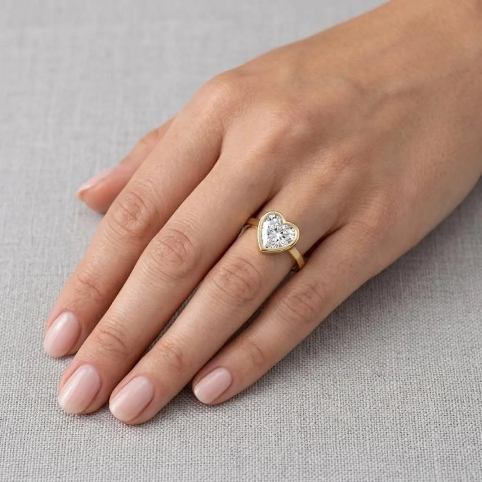 On-hand view of a 3ct heart cut lab grown diamond bezel set solitaire ring in yellow gold.